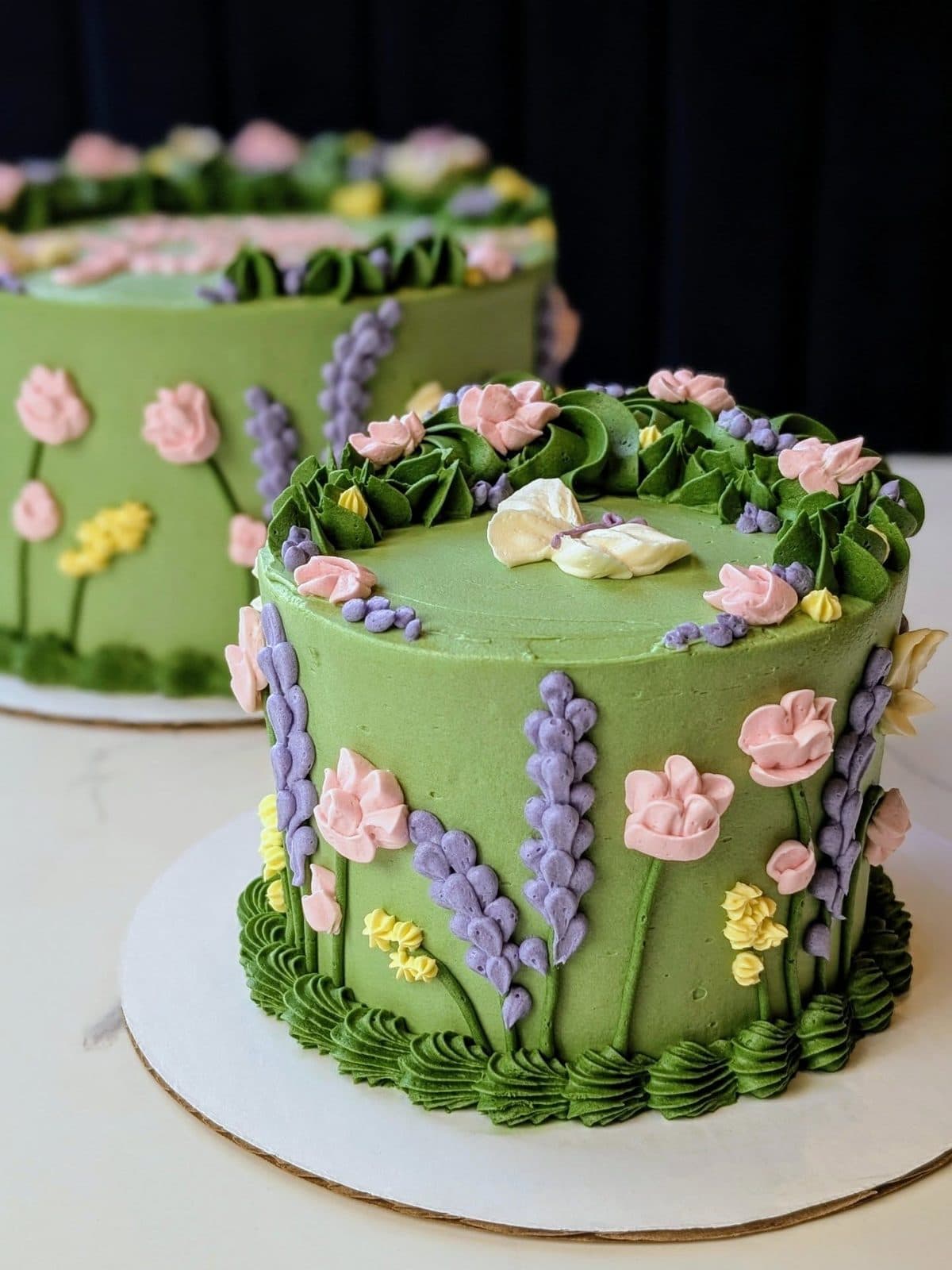 Sage-green buttercream cake covered in piped pink, blue, and white flowers, with one lit candle, on a white plate.